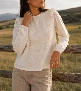 Blusa de color beige con bordados florales en el frente, escote redondo y mangas largas con puños elastizados. Presenta volados decorativos a los lados del pecho.