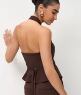 Chaleco de sastrería color marrón, con diseño de frente única y cuello halter. Presenta pinzas laterales que definen la silueta, bolsillos decorativos y ruedo levemente redondeado. En la espalda, cuenta con una cinta ajustable con ojal metálico y tiras largas.
