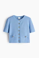 Cárdigan corto de punto acanalado en color celeste, con cuello redondo, manga corta y cierre frontal de botones dorados. Presenta dos bolsillos delanteros decorativos con botón dorado.