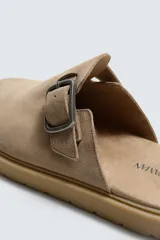 Zueco tipo mule de hombre, color beige topo, fabricado en piel con acabado serraje. Presenta una tira con hebilla metálica cuadrada en el empeine y suela gruesa de goma en color natural contrastante.