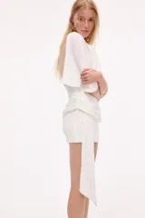 Vestido corto blanco con mangas cortas amplias y cuello recto. Presenta un fajín drapeado de lentejuelas plateadas a la cintura con un lazo largo lateral.
