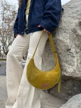 Cartera tipo hobo de cuero gamuzado color verde oliva, con cierre superior y correa ajustable. Presenta una etiqueta de cuero con el logo de la marca en el frente.