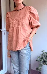 Blusa de lino color salmón con cuello alto, mangas abullonadas y detalle de alforzas en el frente. Presenta terminaciones con puntilla ondulada negra y cierre posterior con botones a la vista.