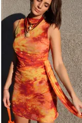 Vestido corto sin mangas con cuello redondo, confeccionado en microtul con un estampado en tonos cálidos como rojo, naranja y amarillo que simula un efecto tie-dye. Presenta un diseño ajustado al cuerpo con detalle de drapeado lateral en la cintura.