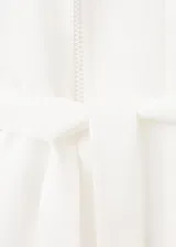Vestido midi camisero de lino color blanco, sin mangas, con cuello solapa, cierre frontal de botones y cinturón ajustable del mismo género. Cuenta con detalles de costura calada en el frente.