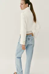 Chaqueta corta blanca de estilo militar, con cuello subido, manga larga y trabillas desflecadas en los hombros. Cuenta con cierre frontal cruzado decorado con botones metálicos dorados.