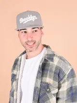 Gorro tipo snapback negro con visera plana y bordado frontal en blanco con la palabra "Brooklyn" en cursiva.