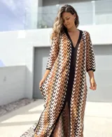 Vestido largo de tejido de punto con diseño geométrico en tonos tierra, negro y blanco. Presenta escote en V profundo, mangas tres cuartos y tajos laterales en la falda.