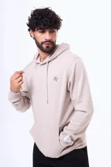 Buzo tipo canguro con capucha y cordones ajustables, confeccionado en felpa color beige. Presenta un pequeño logo bordado en el pecho y una etiqueta decorativa en el puño de la manga.