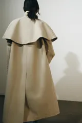 Gabardina larga de corte recto y diseño minimalista en color beige, con cuello solapa, manga larga y cierre frontal con botones. Cuenta con una capa desmontable en el mismo tejido y una solapa decorativa en el pecho.