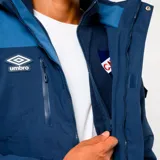Parka técnica de invierno Umbro del Club Nacional de Football, en tonos azul petróleo y azul más claro. Cuenta con capucha ajustable, cierre frontal con solapa, bolsillos frontales con tapa y logo de la marca junto al escudo del club en el pecho.