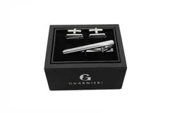 Set de gemelos y sujetacorbata plateados con detalles en negro, presentados en caja negra con logo de la marca.