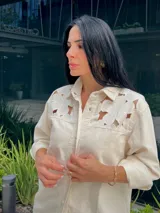 Camisa color off-white con detalles calados en forma de flores en los hombros y cuello. Tiene cierre frontal con botones a presión y cordón decorativo.