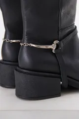 Bota de cuero negra con caña media, taco cuadrado y detalle de correa con pieza metálica plateada alrededor del tobillo.