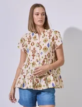 Blusa color crema con estampado tribal en tonos marrón, verde y azul. Tiene cuello a la base, escote en V, abertura central con botones, corte en la cintura con volados y ruedo con volados.
