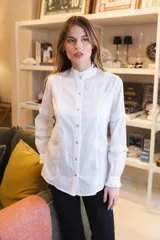 Camisa blanca de algodón con cuello alto tipo mao con volados, cierre frontal de botones y mangas largas con detalles de volados horizontales y puños con volados.