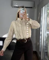 Camisa blanca de estilo romántico con cuello alto, cierre frontal de botones y detalles de encaje y alforzas verticales. Presenta mangas largas con puños decorados con encaje.
