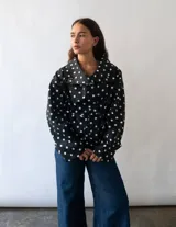 Campera de algodón con estampado de lunares blancos sobre fondo negro. Presenta cuello alto, cierre frontal con botones metálicos y cinturón ajustable en la cintura. Posee bolsillos laterales y un diseño versátil que permite llevarla entallada o con silueta holgada.