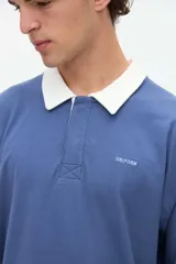 Remera polo de manga larga azul con cuello camisero blanco y cierre frontal de botones ocultos. Presenta un pequeño bordado con el texto "uniform" en el pecho y calce relajado.