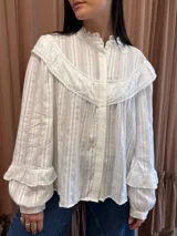 Camisa blanca de manga larga con textura a rayas verticales, canesú con volados y detalles de encaje en el pecho y el cuello alto. Cuenta con cierre frontal de botones ocultos y mangas abullonadas con volados en los puños.
