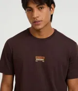 Remera de algodón peruano color marrón oscuro, corte comfort, con cuello redondo y mangas cortas. Presenta un pequeño parche rectangular estampado en el pecho con texto en tonos naranja y blanco.