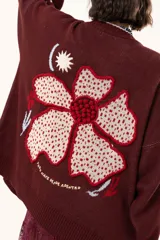 Saco de punto color vino con diseño holgado, cuello redondo y mangas largas. Presenta un gran bordado central en la espalda con forma de flor en tonos crema y rojo, acompañado de un pequeño bordado de sol y una frase bordada en la parte inferior.