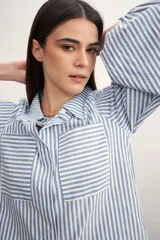 Camisa a rayas verticales color beige y blanco, con cuello clásico, cierre frontal con botón dorado, mangas largas abullonadas con puños elásticos, dos bolsillos frontales y corte holgado.