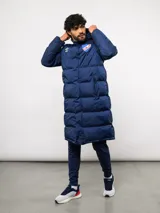 Parka larga acolchada color azul marino con capucha, cierre frontal con botones a presión y bolsillos laterales. Presenta el logo de Umbro y el escudo del Club Nacional de Football bordados en el pecho.