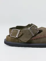 Slipper tipo guillermina confeccionada en gamuza color marrón, con plataforma de goma negra y doble hebilla ajustable. Presenta un pespunte decorativo en el borde inferior y cuenta con interior recubierto en cuero.