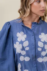 Camisa azul de calce relajado con bordado floral blanco en el frente. Presenta mangas largas amplias con ligero fruncido en el hombro y cierre frontal con botones.