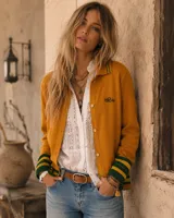 Cárdigan de punto de lana merino color mostaza con cuello tipo polo, cierre frontal de botones y detalles de rayas verdes y amarillas en los puños y el borde inferior. Cuenta con un bordado sutil en el pecho.