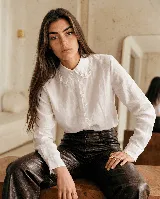 Camisa blanca de lino con cuello tipo Peter Pan amplio adornado con puntilla bordada, cierre frontal oculto y mangas largas con puños rectos.