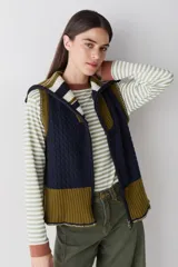 Chaleco de lana con cierre frontal completo, cuello alto y diseño bicolor en tonos marrón y celeste. Presenta una combinación de tejido trenzado y acanalado, con un corte asimétrico más largo en la parte posterior y aberturas laterales.