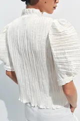 Blusa de cuello redondo con detalle de volados, manga corta abullonada con terminación elástica y cierre frontal con botones. Presenta un diseño de tejido plisado en toda la prenda.