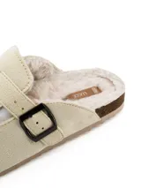 Pantufla tipo zueco en color beige, confeccionada en material tipo gamuza con interior forrado en piel sintética. Cuenta con una tira ajustable en el empeine con hebilla metálica lateral.