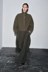 Chaqueta corta de tejido grueso color verde oliva, con cuello alto y cierre frontal completo de cremallera plateada. Presenta un corte amplio y mangas largas caídas.