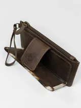 Billetera porta celular confeccionada en cuero marrón con detalle de pelo vacuno con estampado animal print tipo vaca en tonos blancos, marrones y negros. Cuenta con correa de muñeca y cierre superior.