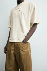 Camiseta de manga corta y cuello redondo, color crema o blanco roto, con un parche texturizado en el pecho que simula barras verticales.