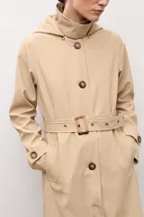 Trench clásico color beige, con cuello alto, capucha desmontable, cierre frontal de botones marrones, bolsillos ribeteados y cinturón ajustable con hebilla.