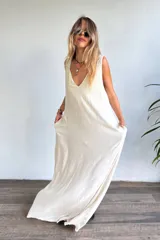 Vestido largo sin mangas, color natural/crudo, con escote en V profundo y corte holgado. Presenta aberturas laterales en la falda.