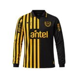 Camiseta de fútbol Puma del Club Atlético Peñarol, edición aniversario manga larga. Diseño con mitad derecha a rayas verticales amarillas y negras, y mitad izquierda negra con escudo retro "CUR" en dorado. Cuenta con cuello tipo polo negro con detalles amarillos y sponsors impresos en el frente y mangas.