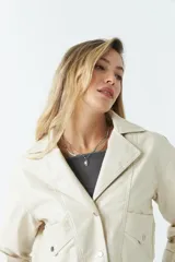Campera de cuero sintético en color crema, con diseño ajustado al cuerpo y largo hasta la cintura. Presenta cierre frontal con botones, bolsillos con solapa y detalles de cierres metálicos en los puños.