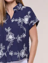 Blusa azul marino de corte amplio en tejido de puro algodón, con cuello camisero abierto en V y detalle de bordado floral en color blanco en toda la superficie. Cuenta con mangas cortas de hombro caído con puño doblado fijo y ruedo curvo con espalda más larga.