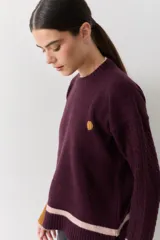 Buzo de punto color crudo con mangas de textura trenzada, detalles en contraste en los puños y franjas horizontales en el ruedo. Presenta cuello redondo y un pequeño bordado en el pecho.
