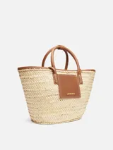 Bolso tipo canasto tejido a mano en fibra vegetal con detalles de cuero marrón, asas cortas enrolladas y logotipo Jacquemus en letras doradas en el frente.