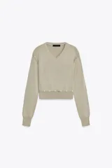 Jersey de punto color beige claro, con cuello en V y manga larga. Presenta detalles de botones decorativos en los hombros y en el ruedo inferior, sugiriendo un diseño desmontable o con aberturas ajustables.