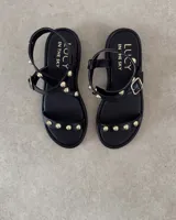 Sandalias planas de cuero negro con tiras decoradas con tachas metálicas doradas. Presentan una tira frontal sobre los dedos y una tira ajustable con hebilla alrededor del tobillo.