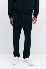 Pantalón jogger negro de corte regular fit, confeccionado en tejido suave con mezcla de algodón. Presenta pliegues cosidos en la parte delantera, cintura elástica con cordón ajustable, bolsillos laterales y un bolsillo insertado en la parte trasera.