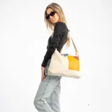 Bolso bandolera de lona color beige, con correa ajustable y diseño de parches en tonos amarillo y marrón en el frente. Cuenta con un detalle de etiqueta azul en la parte superior.