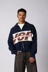 Campera tejida de punto con cierre frontal completo, cuello tipo polo y diseño de bloques de color azul marino, blanco y marrón con estampa gráfica central. Presenta puños y ruedo acanalados.